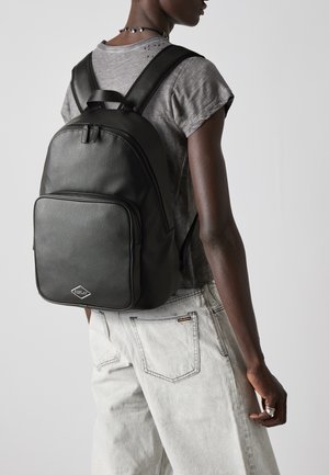 Zaino in pelle nera con una texture liscia, dotato di una tasca frontale con zip e di un logo a diamante. Indossato sulla schiena, abbinato a un abbigliamento grigio.