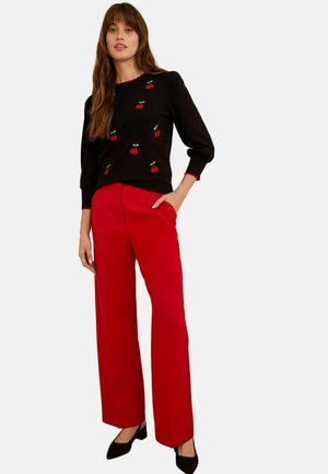 Maglione nero con ricamo di ciliege e bordi rossi, abbinato a pantaloni larghi rossi brillanti e tacchi a punta neri.