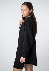 Black oversized shirt made of textured fabric, featuring long sleeves, a collar, and a curved hem. Paired with black shorts.