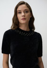 Czarny sweter z krótkim rękawem wykonany z miękkiego, puszystego materiału. Posiada wykończenie z koralikami na dekolcie z rozproszonymi okrągłymi akcentami.