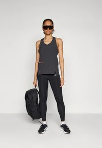 Svart atletisk linne med v-ringning, kombinerat med svarta leggings. Personen håller en svart ryggsäck. Mörka solglasögon kompletterar looken.