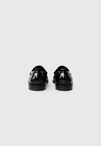 Zapatos de charol negros con una textura suave, tacón bajo y tirador trasero. Presentan un diseño clásico con sutiles detalles de costura.