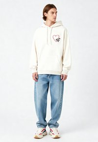 Felpa bianca con un cuore rosso e un design grafico nero sul petto, tasca a marsupio, polsini a coste, abbinata a jeans azzurri e sneakers.