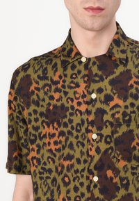 Camisa de manga corta con estampado de leopardo en base verde, manchas marrones y naranjas, con botones al frente y un bolsillo en el pecho. Tejido texturizado.