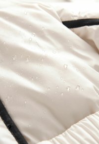 Helle, wasserfeste Stoff mit kleinen Wassertropfen, glatte Textur, geraffter Saum und kontrastierender schwarzer Besatz entlang der Kanten.