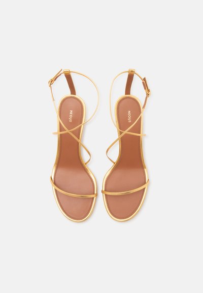 Paire de sandales plates dorées avec fines brides à la cheville et semelles marron vues de dessus sur fond blanc.