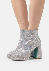 Silver rhinestone-covered ankle boots with a square toe and a chunky teal heel. Smooth texture and shiny surface create a sparkling effect.