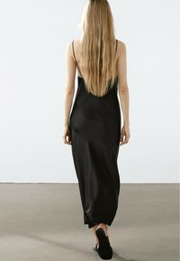 Abito maxi in satin nero con spalline sottili, schiena bassa e silhouette aderente. Sembra liscio al tatto con un design fluido.