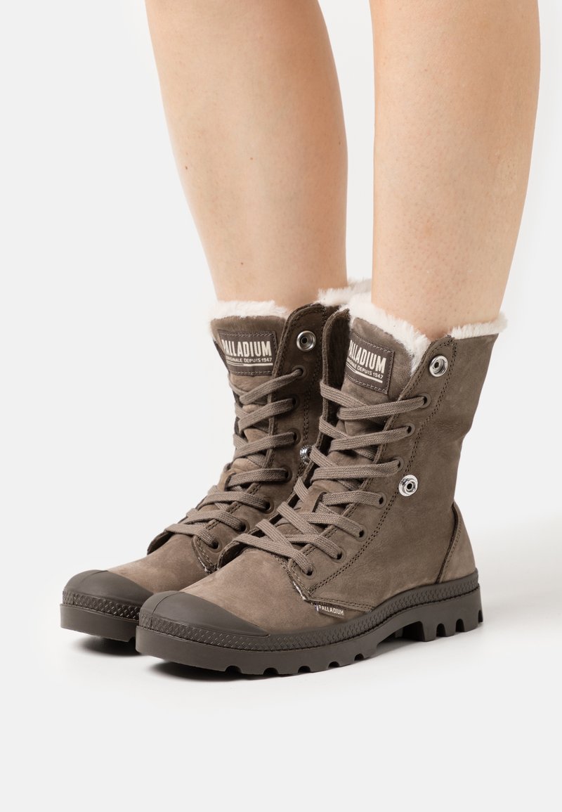 Bottes d'hiver marron à lacets avec doublure en fourrure blanche portées sur des jambes nues sur fond blanc.