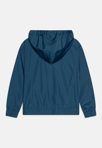 Marineblå, lett vindtett jakke med hette, elastiske mansjetter og kant. Glatt tekstur og minimalistisk design. Bakvinkel kun.