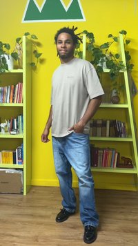 Beige short-sleeve shirt, light blue jeans, and black shoes. Background features green plant shelves and a bright yellow wall.