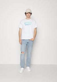 Witte katoenen t-shirt met pastel Levi's logo, gecombineerd met lichtblauwe versleten jeans en witte sneakers, afgewerkt met een beige pet.