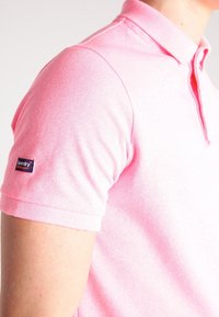 Koszulka polo w jasnoróżowym kolorze, uszyta z bawełny o teksturowanym wykończeniu, z kołnierzykiem, krótkimi rękawami i granatową naszywką z logo na lewym rękawie.