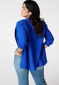 Blazer bleu avec manches retroussées, coupe décontractée et détails d'évent au dos. Associé à un jean en denim bleu clair. Texture lisse, design ajusté.