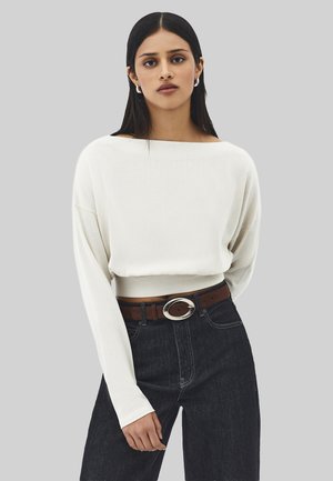 Vrouw die een wit langarmig topje met boothals draagt, donkere high-waist jeans en een bruine riem met een zilveren ovale gesp.