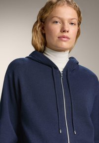 Námornícky s kapucňou, zipsový sveter vyrobený z mäkkej pleteniny, má rebrované akcenty a hladký zips. Model má pod ním biele rolák.