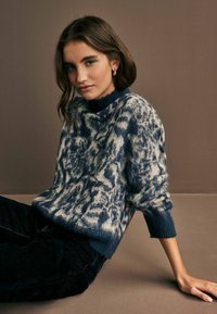Gestreifter Pullover in Blau und Weiß mit einem hohen Kragen und abstraktem Muster. Weiche Textur mit gerippten Bündchen. Kombiniert mit dunklen Jeans.