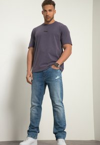 Camiseta de algodón púrpura con un acento negro, combinada con jeans desgastados de color azul claro y zapatillas blancas, de pie frente a un fondo neutro.
