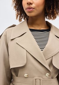 Γυναίκα με μπεζ παλτό τύπου trench πάνω από μαύρο και άσπρο ριγέ τοπ, δείχνοντας το γιακά, τα κουμπιά και τις λεπτομέρειες της ζώνης του παλτού.