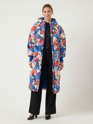 Woman wearing a long, colorful floral puffer coat with a hood, paired with black wide-leg pants and black heeled shoes, standing against a white background.