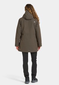 Kobieta z długimi kręconymi włosami, ubrana w brązową kurtkę z kapturem, czarne spodnie i czarne buty trekkingowe, stojąca tyłem na jednolitym tle.