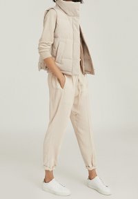 Jasnobeżowa pikowana kamizelka z wysokim kołnierzem, zestawiona z dopasowanymi, luźnymi spodniami i białymi sneakersami. Gładka tekstura, minimalistyczny design.