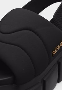 Μαύρο επενδεδυμένο σανδάλι με χοντρή σόλα, ανοιχτό δάχτυλο, ελαστικό λουράκι στο τακούνι και χρυσό μεταλλικό λογότυπο της μάρκας στο πλάι.