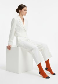 Bela, po meri krojena blazer jakna prek ujemajočih skrajšanih hlač, kombiniranih z oranžnimi nogavicami s temnim poudarkom na prstih. Model sedi na belem kocki.