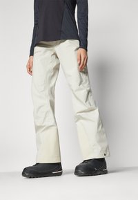 Pantalons de ski imperméables de couleur claire avec des jambes inférieures renforcées et un logo. Associés à des bottes de neige noires avec des accents gris.