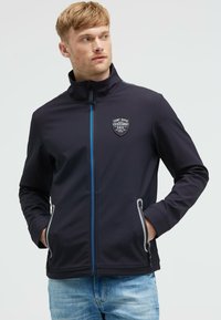 Chaqueta de cremallera navy con cuello alto, que presenta un parche de logo y acentos grises. Material suave con dos bolsillos laterales.