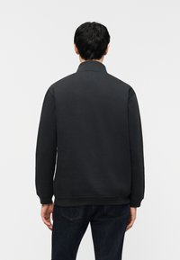 Άνδρας που φοράει απλό μαύρο φούτερ και σκούρο τζιν, βλεπόμενος από πίσω μπροστά σε απλό λευκό φόντο.