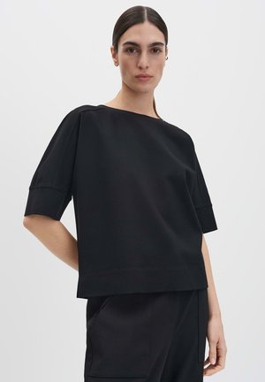 Haut noir à manches courtes en tissu doux et lisse, de forme carrée avec un décolleté large. Design minimaliste sans motifs ni accents visibles.