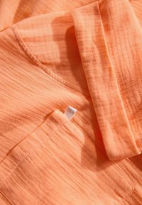 Baumwollhemd in hellorange mit strukturiertem Finish. Verfügt über eine Tasche und ein beschriftetes Etikett. Der zerknitterte Look verstärkt die Textur des Stoffes.