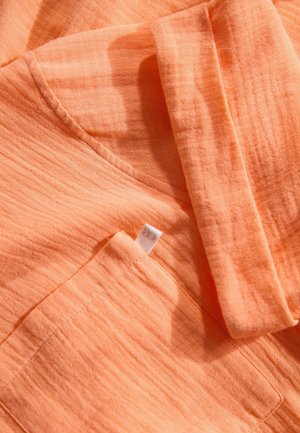 Katoenen shirt in lichtoranje met een gestructureerde afwerking. Beschikt over een pocket en een label. Kreukelige uitstraling versterkt de textuur van de stof.