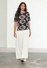 Black top with a textured beige shell pattern, short sleeves pairs with cream wide-leg pants; model stands on a neutral floor.
