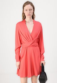 Coral wrap dress with long sleeves, deep V-neck, and gathered waist. Smooth fabric, flared skirt, and a hand-held black bag.