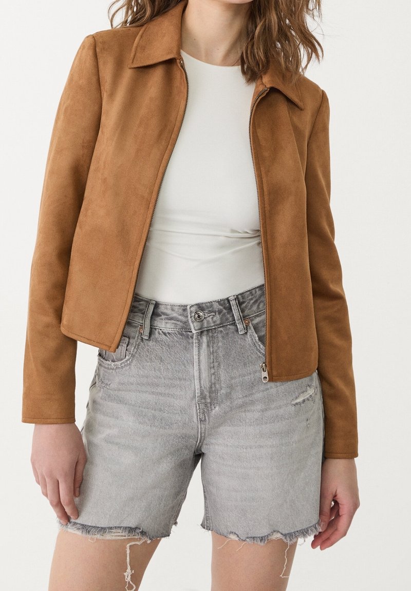 Ruskea tekoturkispuku, jossa on kauluspäällinen ja etuvetoketju; yhdistetty harmaisiin kuluneisiin denim-shorteihin, joissa on hapsut.