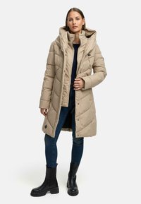 Beige polstret lang frakk med høy krage og hette, med diamantkviling og snapknapper, kombinert med mørke jeans og svarte ankelstøvler.