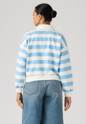 Femme aux cheveux foncés en chignon, portant une chemise à manches longues rayée bleu et blanc et un jean large bleu clair, vue de dos.
