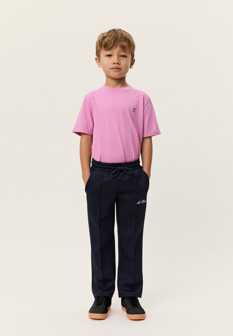 Ροζ βαμβακερό μπλουζάκι με κοντά μανίκια και μικρό λογότυπο στο στήθος, συνδυασμένο με ναυτικά παντελόνια με κορδόνι και λευκή κεντητική διακόσμηση, και μαύρα αθλητικά παπούτσια.