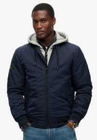 Marine bomberjack met een grijze hooded voering, ritsluiting, zijzakken, geribde manchetten en een gladde textuur. Gecombineerd met denim jeans.