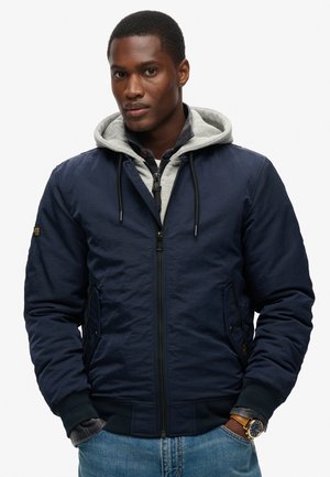 Navy bomber jacket with a gray hooded lining, zip closure, side pockets, ribbed cuffs, and a smooth texture. Paired with denim jeans.