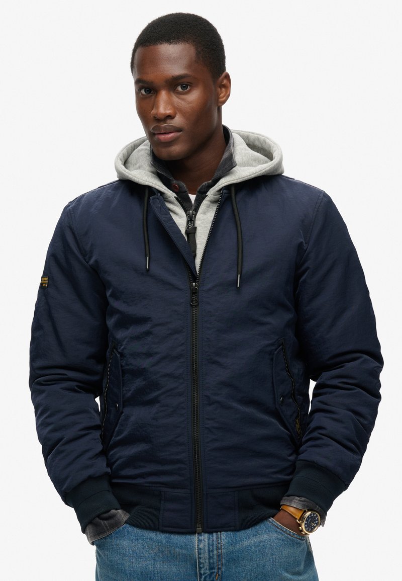 Marine bomberjack met een grijze hooded voering, ritsluiting, zijzakken, geribde manchetten en een gladde textuur. Gecombineerd met denim jeans.