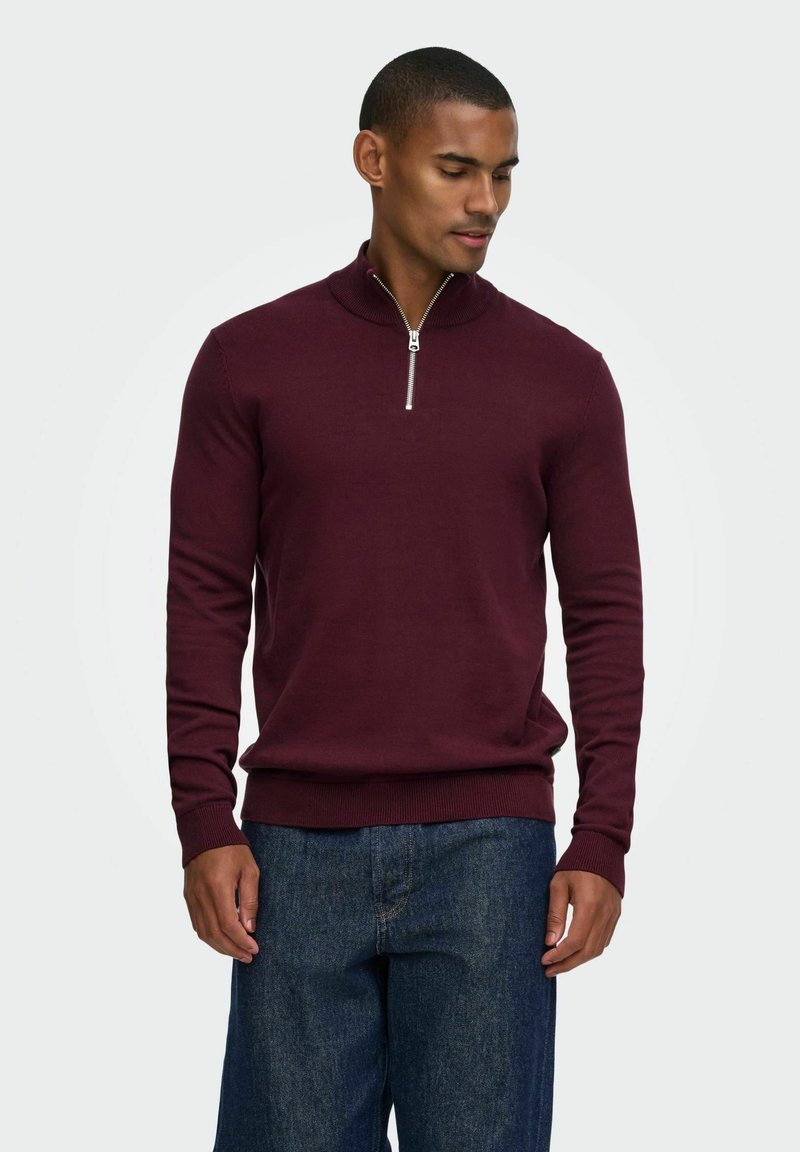 Homme portant un pull bordeaux à manches longues avec une demi-fermeture éclair et un jean bleu, regardant vers le bas à sa gauche sur un fond uni.