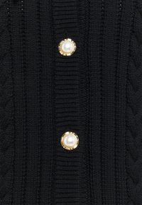 Black knitted cardigan featuring a ribbed texture and two large round buttons with a synthetic pearl center surrounded by a gold rim.