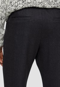 Dunkelgraue Hosen aus strukturiertem Stoff, mit einer Reihe von Gesäßtaschen, und einem teilweise sichtbaren grauen Strickpullover darüber.