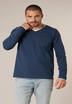 Man met kort haar en baard, gekleed in een marineblauw shirt met lange mouwen en lichte spijkerbroek, die zijn nek aanraakt tegen een effen achtergrond.