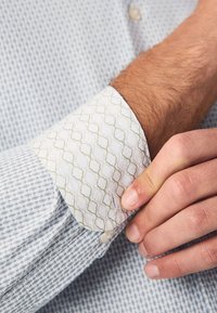 Világoskék mintás ing fehér mandzsettával, amely zöld hullám mintával díszített. Az anyag texturált, szövött megjelenésű.