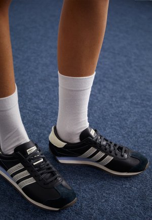 Fekete sportcipők három fehér csíkkal, sima bőr és velúr külsővel, fehér zoknikkal párosítva, kék szőnyegen.