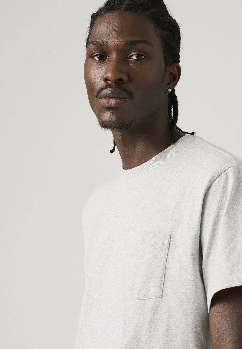 Junger schwarzer Mann mit geflochtenem Haar und Ohrring, trägt ein hellgraues T-Shirt mit kurzen Ärmeln und einer Brusttasche, neutraler Hintergrund.